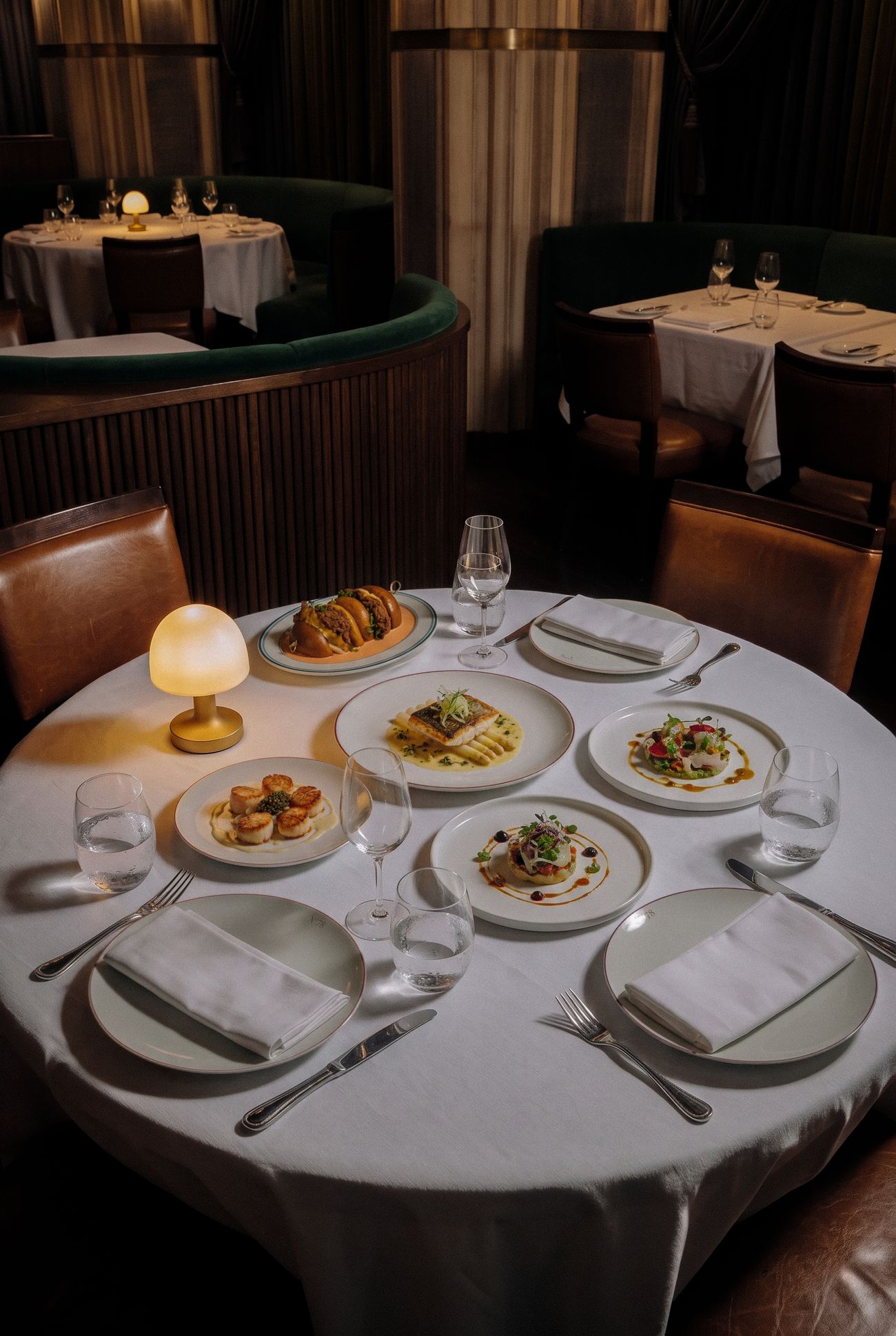 A round table laid with five plates, wine glasses and a glowing lamp