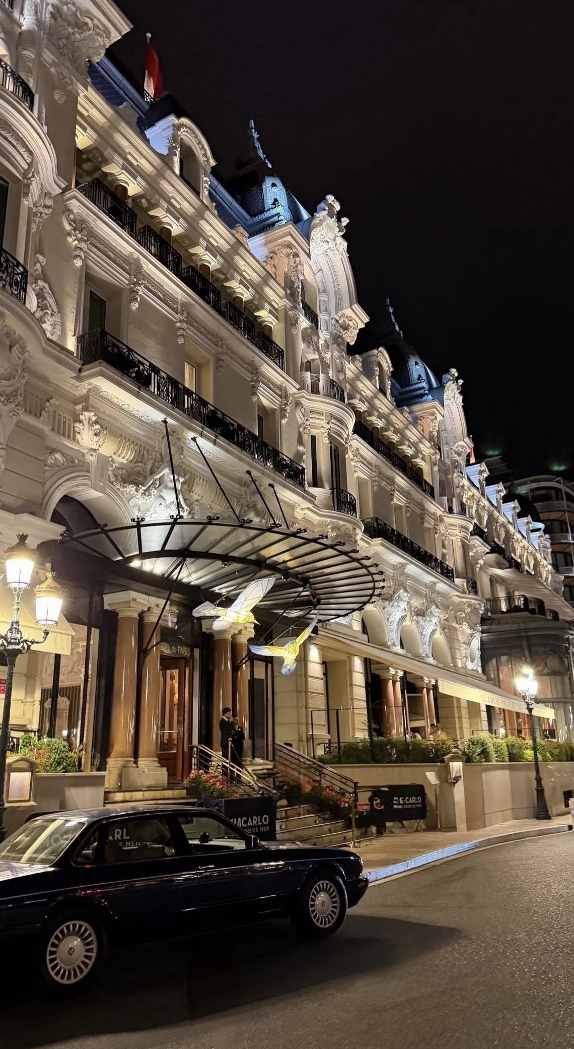 Illuminated stone facade and glass canopy at night, with a dark classic car parked outside
