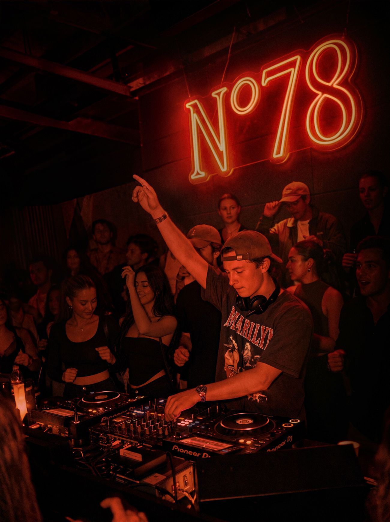A DJ with one arm raised beneath the glowing N°78 neon sign, guests dancing behind him