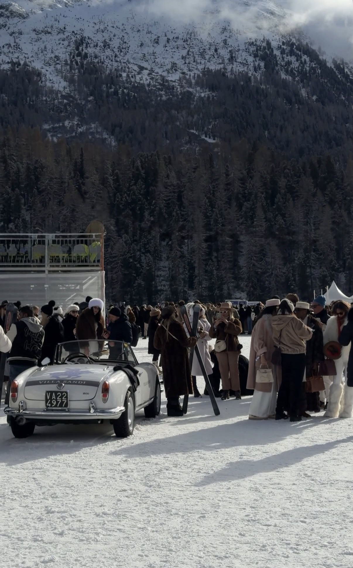 Guests in furs and hats gather around a white vintage convertible on the snow, with a forested mountain behind