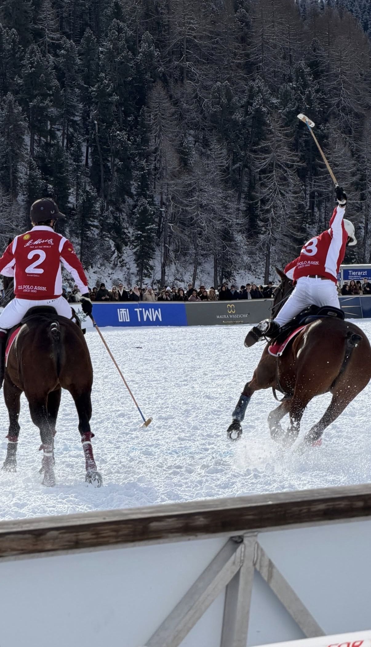 Two polo riders in red and white shirts gallop across the snow, one with his mallet raised