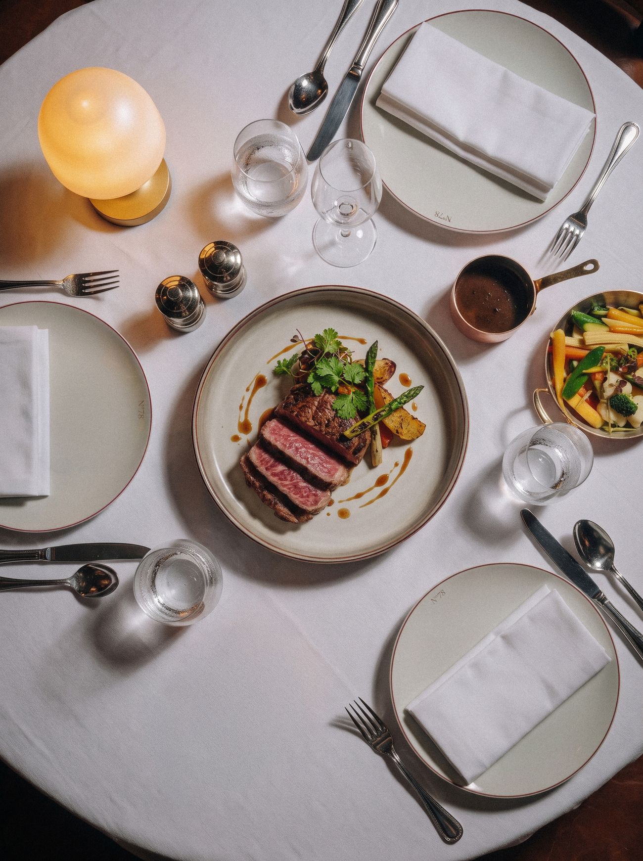 Sliced steak with asparagus on a plate marked N°78, set on a white tablecloth beside a glowing table lamp
